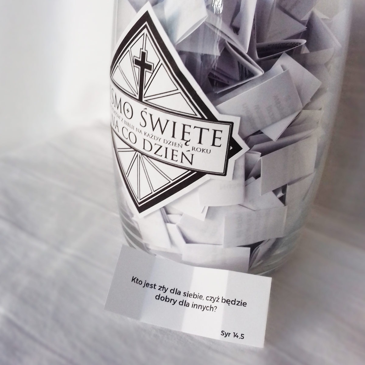 Szklany słoik wypełniony pociętymi kartkami z cytatami biblijnymi, z naklejką z krzyżem i napisem 'Słowo Święte na co dzień' na słoiku oraz pojedyncza kartka z cytatem 'Kto jest zły dla siebie...