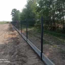 Długi, prosty odcinek ogrodzenia panelowego w kolorze grafitowym, z podmurówką betonową, ciągnący się wzdłuż piaszczystej drogi na tle zieleni drzew i pól.