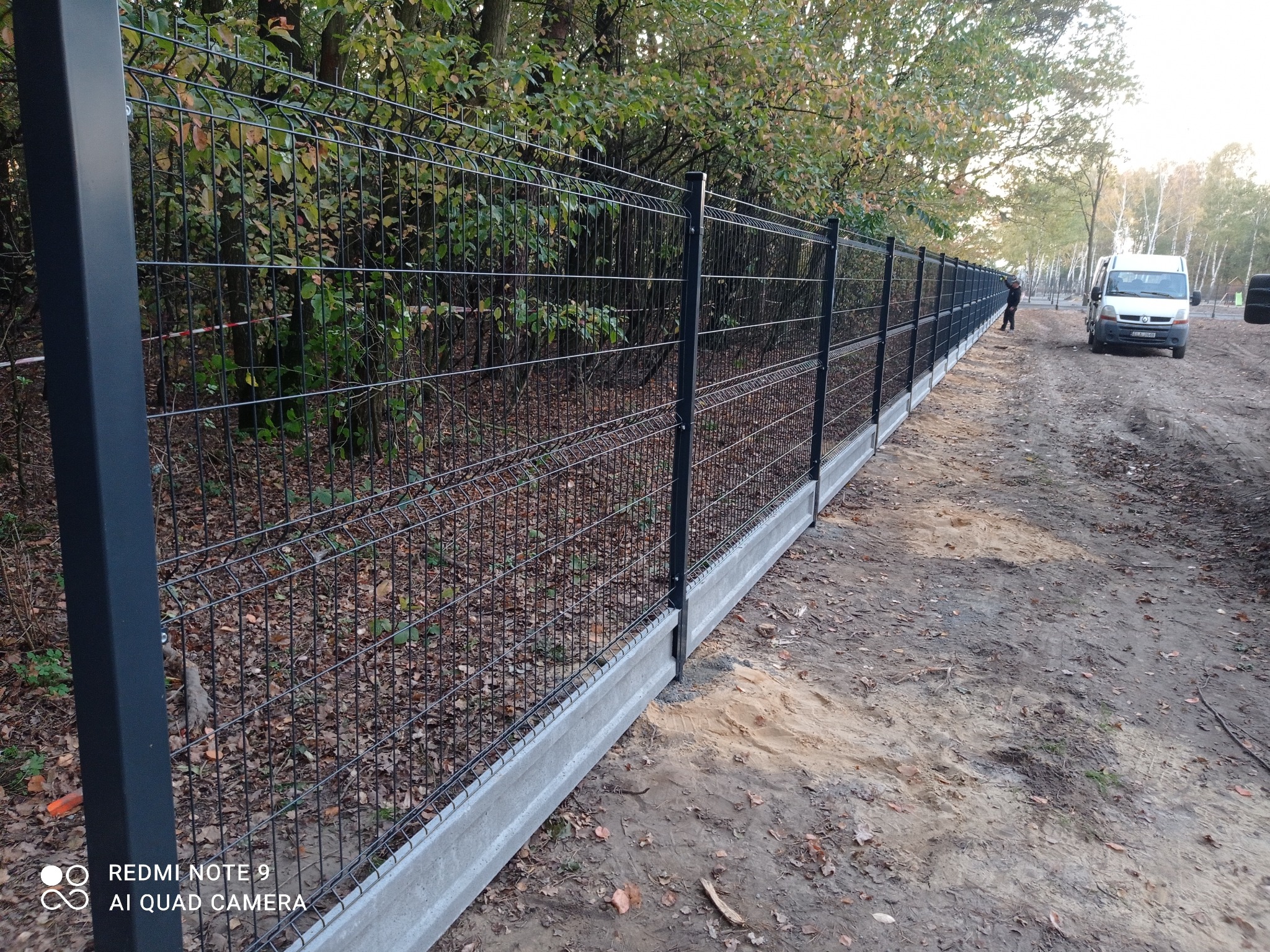 Długi, czarny panel ogrodzeniowy z podmurówką betonową, ciągnący się wzdłuż leśnej drogi. W tle widoczny pracownik i biały samochód dostawczy.
