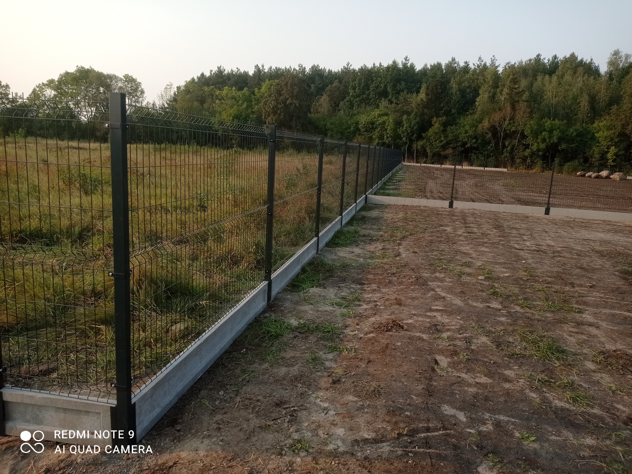 Nowoczesne ogrodzenie panelowe z podmurówką betonową na tle zieleni drzew. Metalowa konstrukcja z siatki tworzy długą linię oddzielającą posesję.