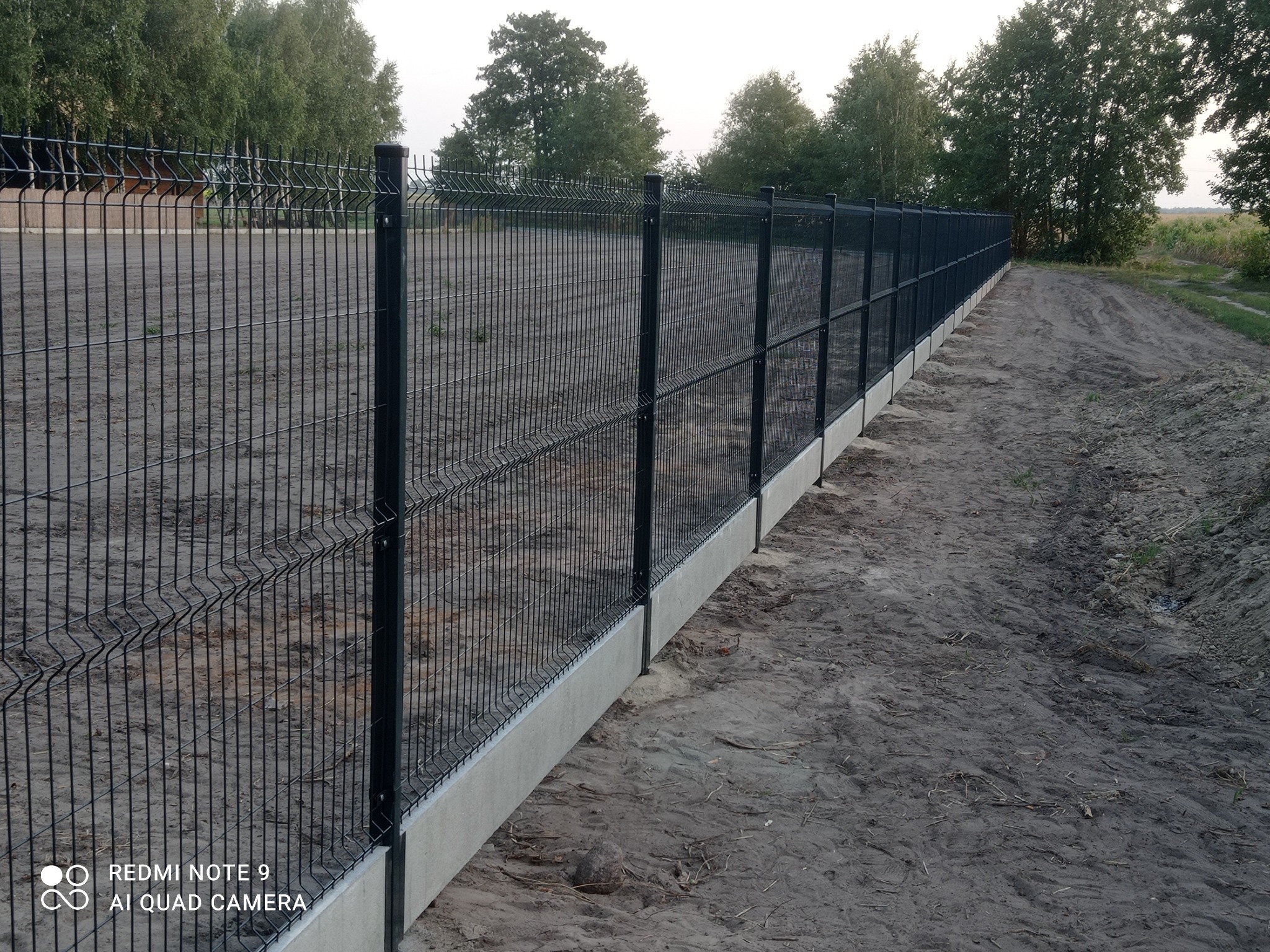 Długi, czarny panel ogrodzeniowy z betonową podmurówką, ciągnący się wzdłuż ziemistej ścieżki, otoczony zielenią drzew w tle. Widok z perspektywy.