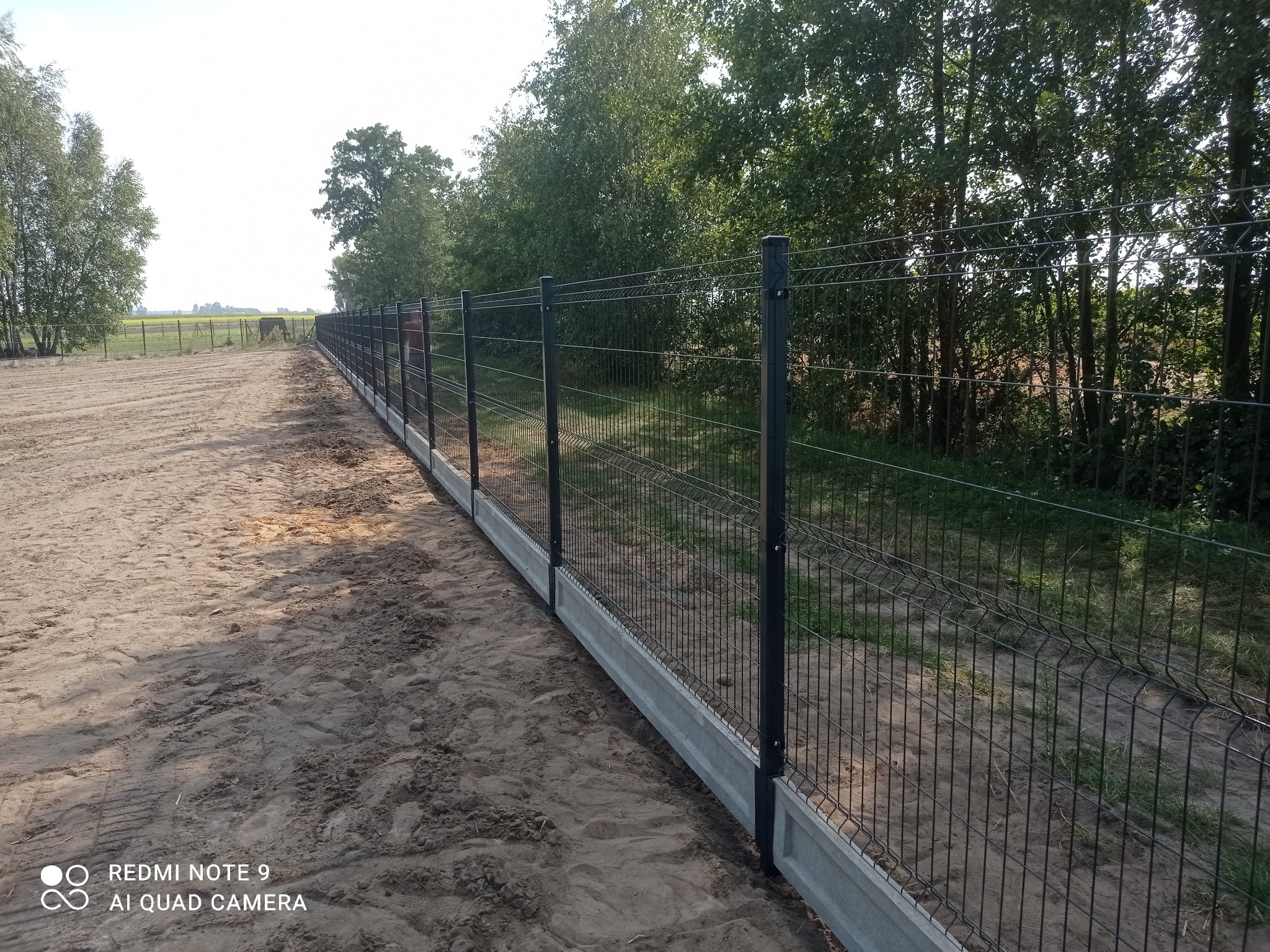 Długi, prosty odcinek ogrodzenia panelowego w kolorze grafitowym, z podmurówką betonową, ciągnący się wzdłuż piaszczystej drogi na tle zieleni drzew i pól.