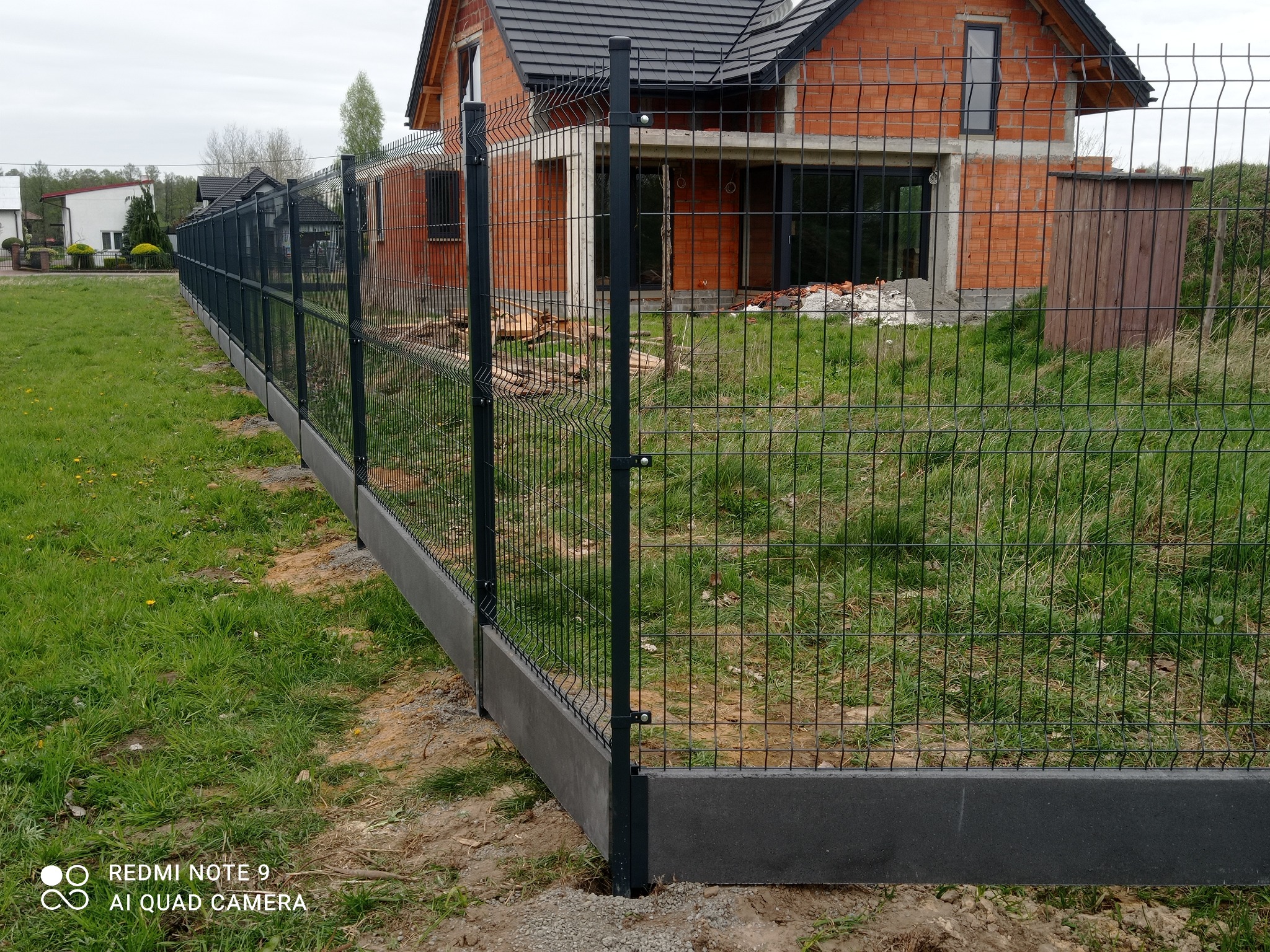 Nowoczesne ogrodzenie panelowe w kolorze grafitowym z podmurówką betonową, otoczone zielenią i nowo budowanym domem z czerwonej cegły w tle.