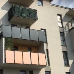 Nowoczesny blok z elewacją w odcieniach szarości i beżu. Balkony z różnymi wypełnieniami: szkło, metalowa siatka i panele w kolorze brzoskwini.