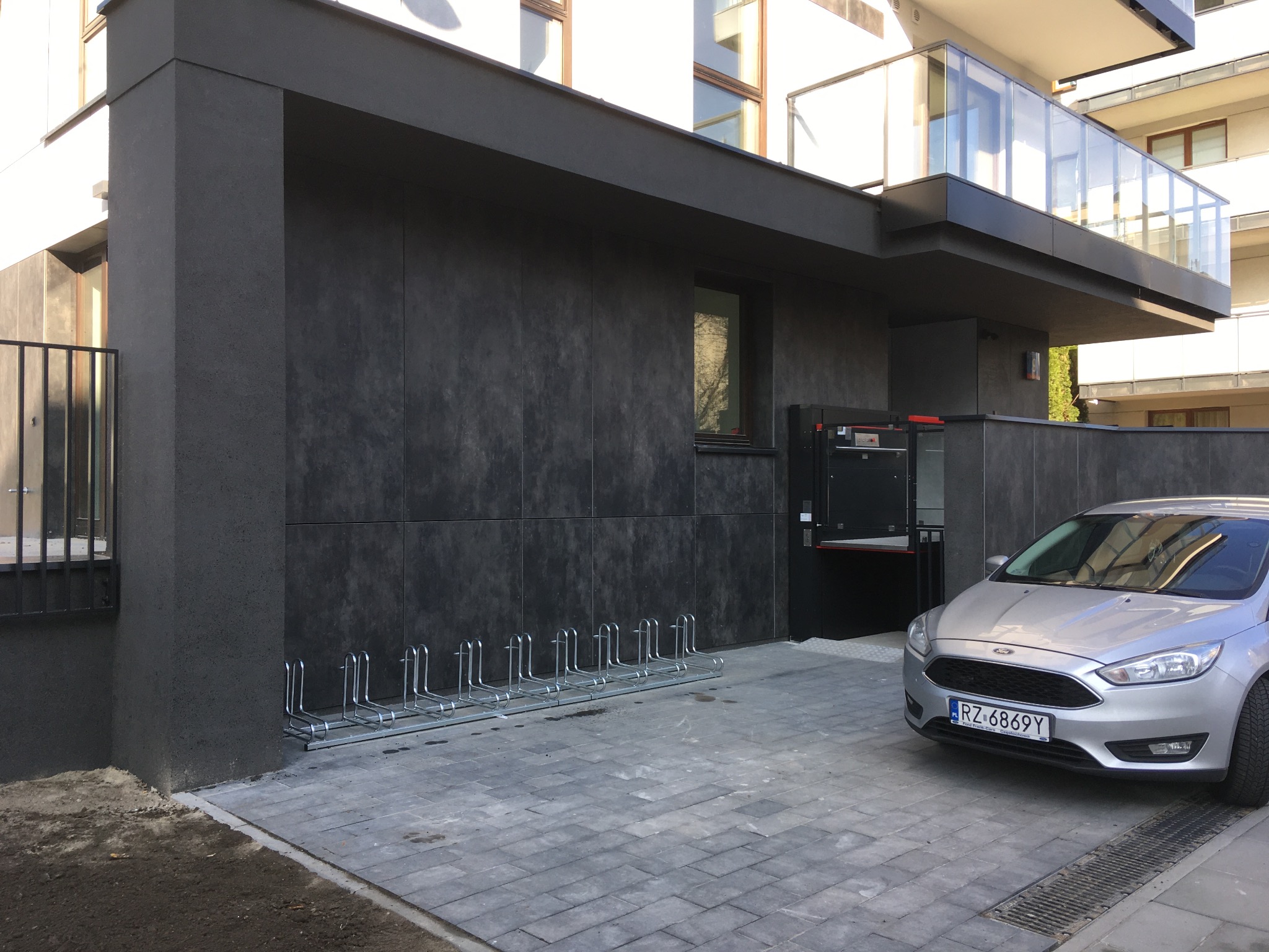 Nowoczesny budynek wielorodzinny z ciemnoszarą elewacją, widoczny fragment parkingu z metalowym stojakiem na rowery i srebrnym samochodem osobowym zaparkowanym obok.