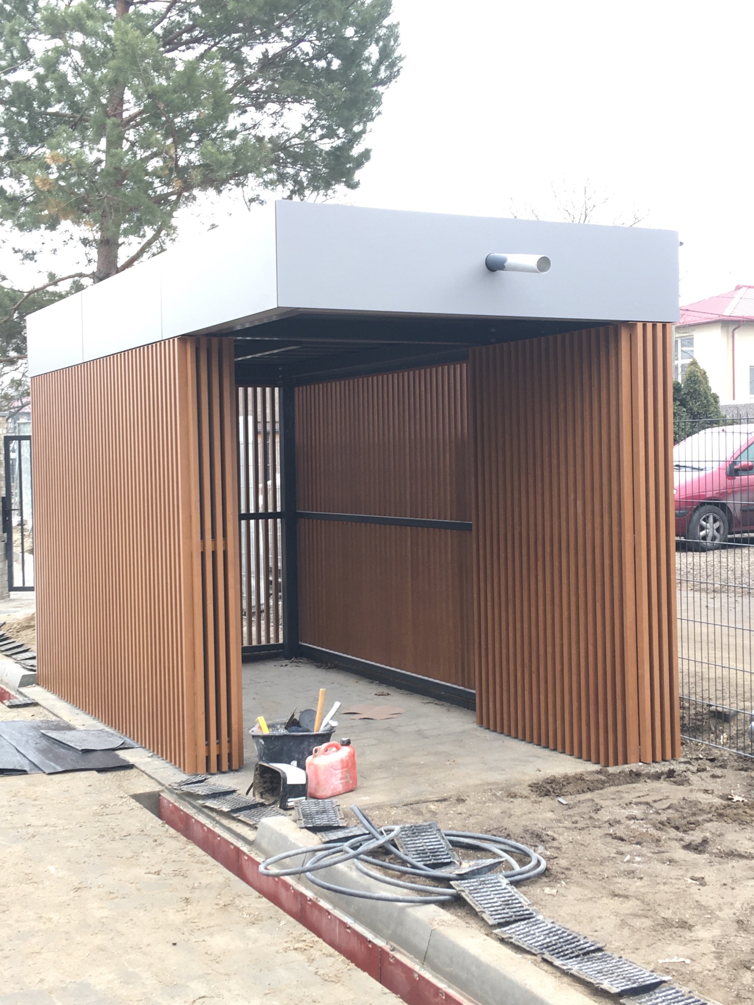 Nowoczesna wiata z drewnopodobnych paneli pionowych w trakcie montażu, widoczne narzędzia, elementy konstrukcyjne i tymczasowe zabezpieczenia na placu budowy.