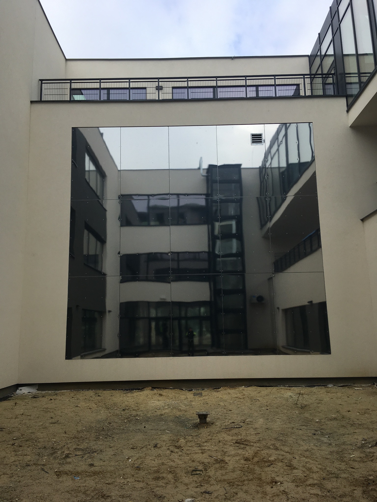 Nowoczesny budynek z elewacją wykonaną z paneli lustrzanych, odbijających sąsiedni budynek, z widocznym placem budowy u podstawy.