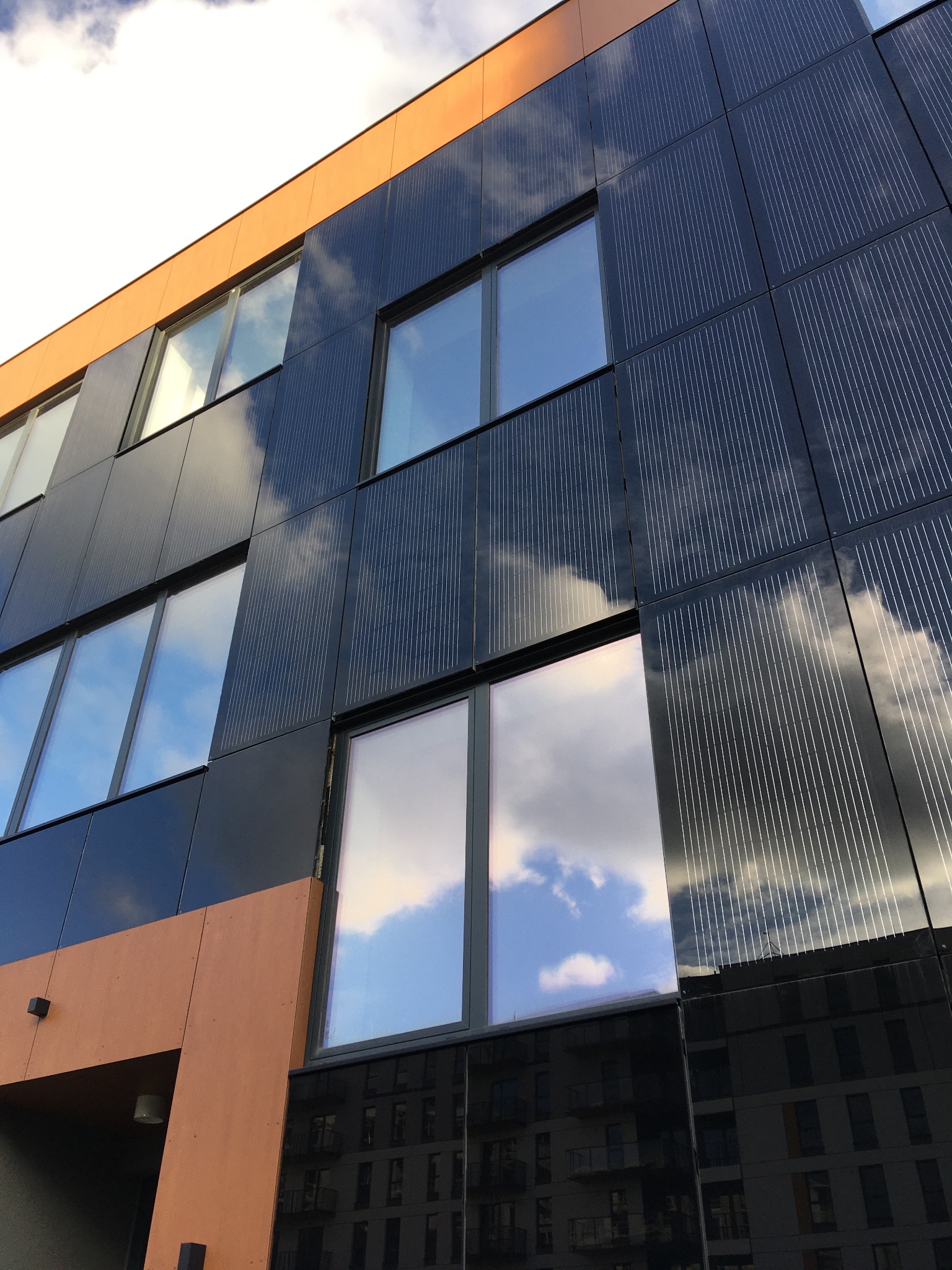 Nowoczesna elewacja budynku w Warszawie z panelami o fakturze drobnych linii, odbijająca błękitne niebo z chmurami w oknach i na powierzchni paneli.