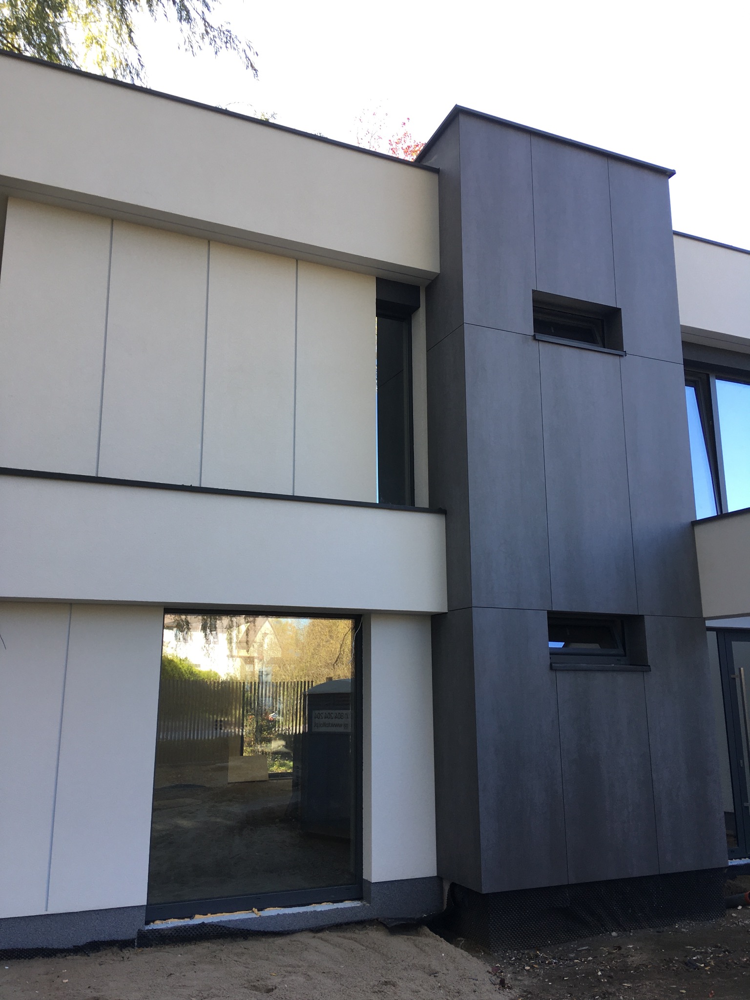 Nowoczesny budynek z elewacją z paneli w odcieniach szarości i bieli, widoczne okna z odbiciem otoczenia, teren wokół budynku w trakcie prac wykończeniowych.