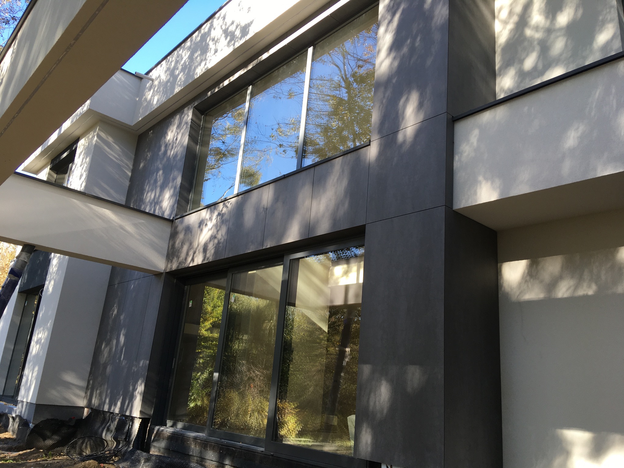 Nowoczesna elewacja budynku z szarymi panelami i dużymi oknami, widoczne prace wykończeniowe przy gruncie.