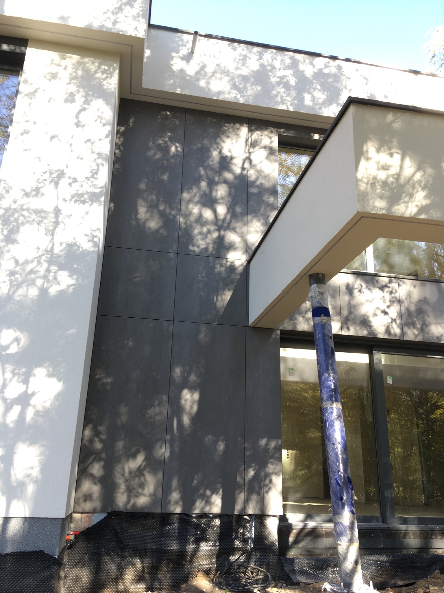 Nowoczesna elewacja budynku z szarymi panelami, białe elementy architektoniczne, widoczne zabezpieczenia budowlane i cień drzew na fasadzie.