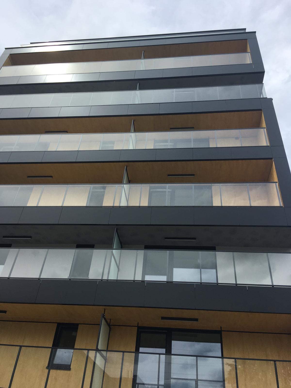 Nowoczesny budynek wielorodzinny z elewacją w odcieniach szarości i beżu, z balkonami z przezroczystymi balustradami, uchwycony z dołu pod pochmurnym niebem.