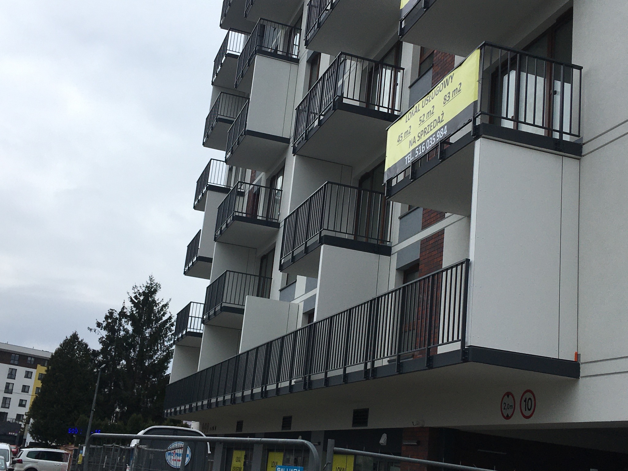 Nowoczesny budynek wielorodzinny z balkonami wykończonymi czarnymi barierkami, na jednym z balkonów baner reklamowy z informacją o lokalach na sprzedaż, widok z dołu na elewację budynku w pochmurny...