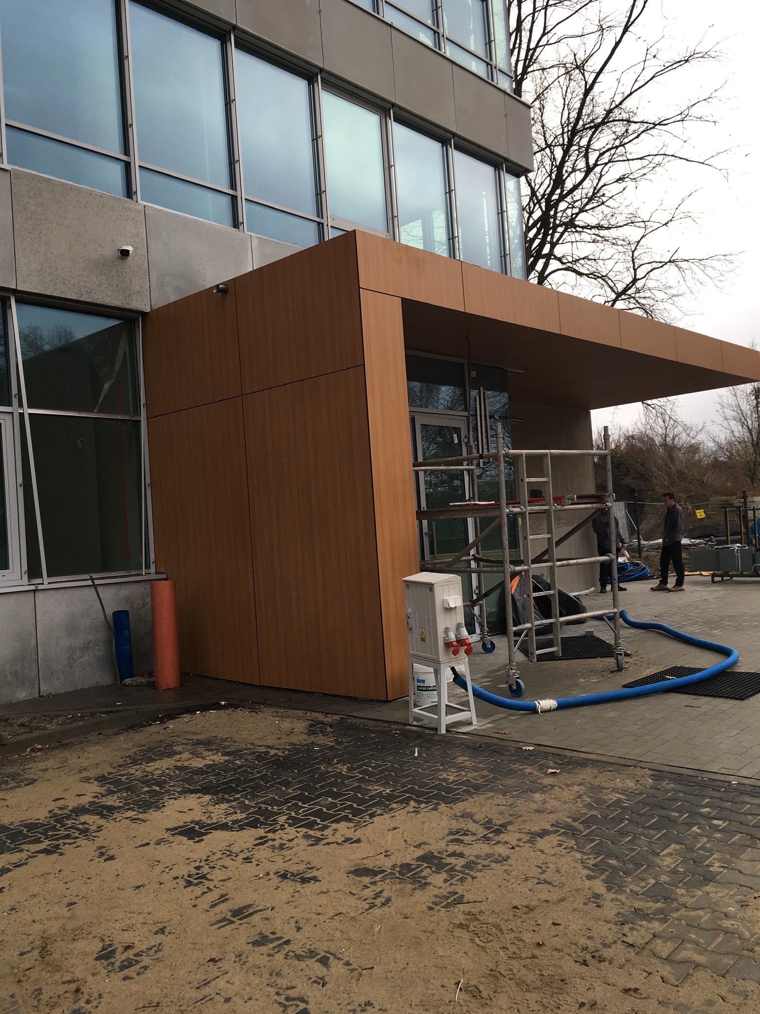 Montaż drewnopodobnej elewacji na budynku biurowym, widoczne rusztowania i ekipa montażowa, fragment elewacji z zamontowanymi oknami.