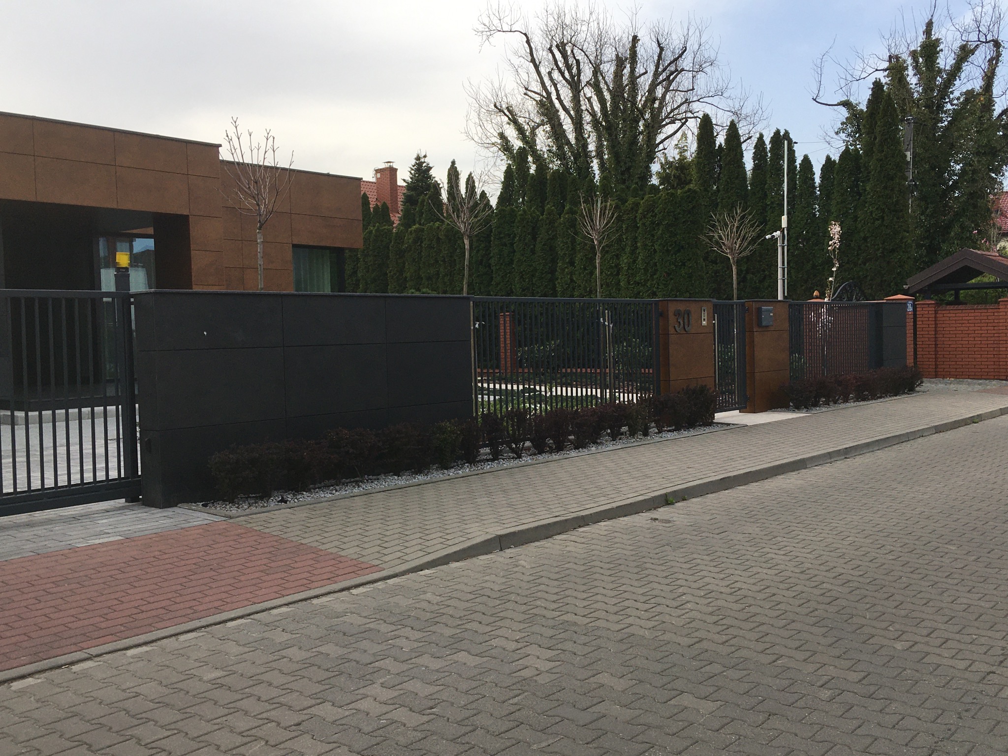 Nowoczesny dom jednorodzinny z brązową elewacją i czarnym ogrodzeniem panelowym, widok z ulicy wyłożonej kostką brukową.