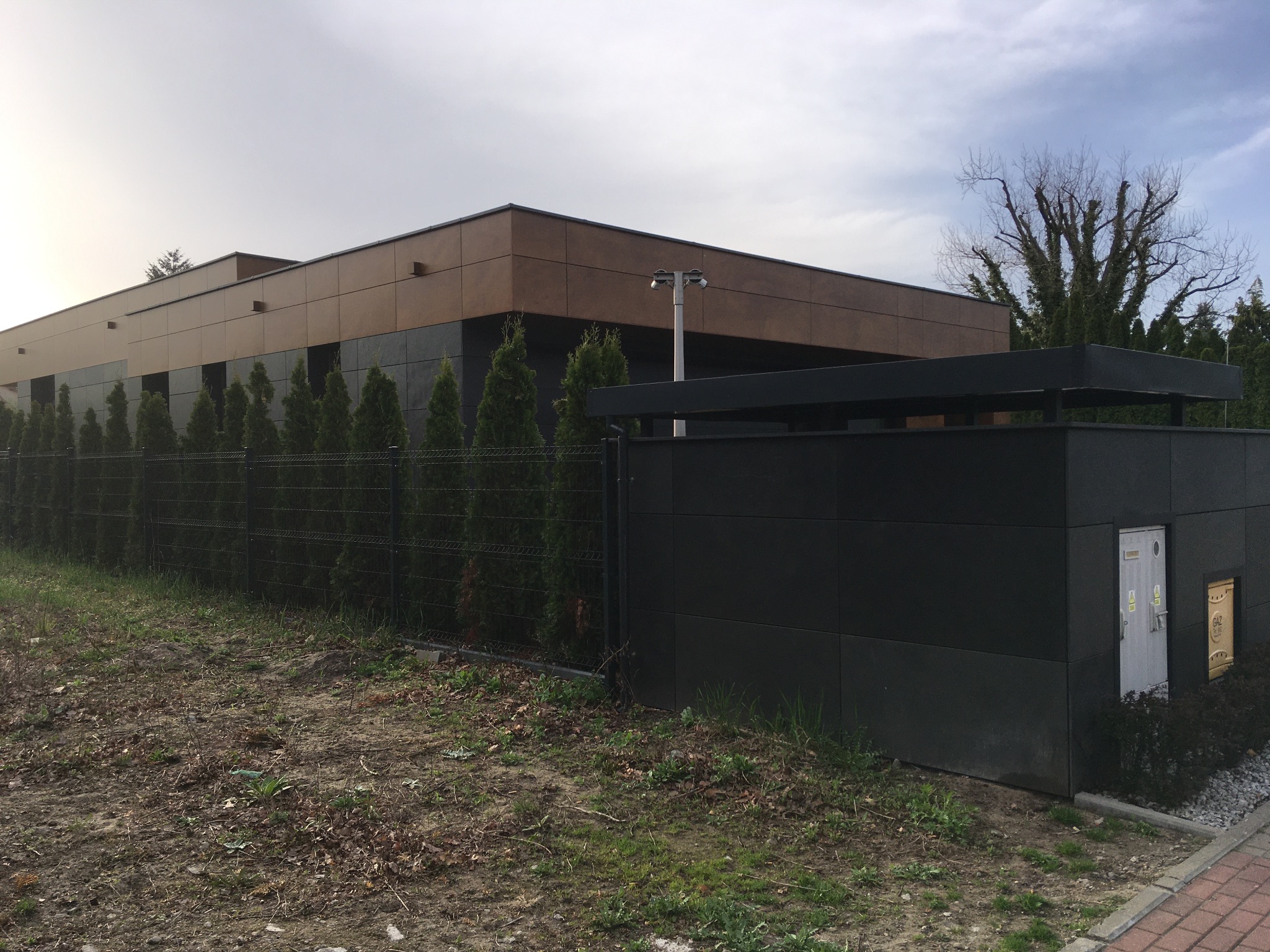 Nowoczesny budynek z elewacją w dwóch odcieniach brązu i czerni, otoczony tujami i metalowym ogrodzeniem, z widocznym słupem oświetleniowym i przyłączem gazowym.