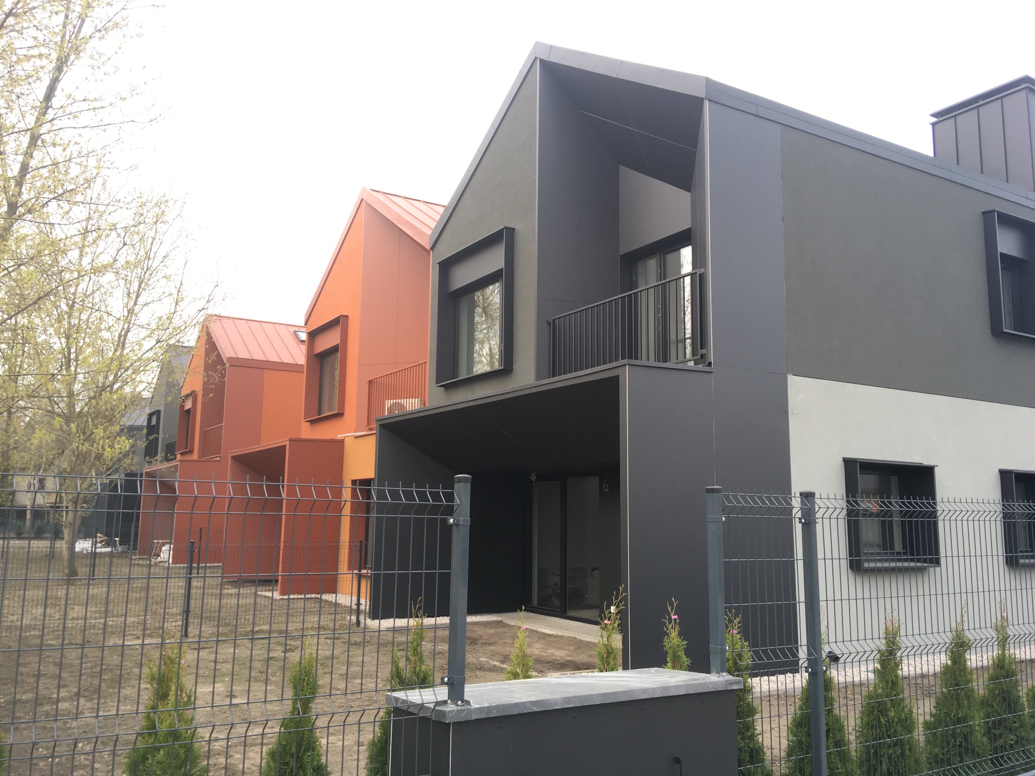 Nowoczesne domy szeregowe z elewacją w odcieniach szarości i pomarańczy, ogrodzone metalową siatką. Widok z zewnątrz na osiedle domów.