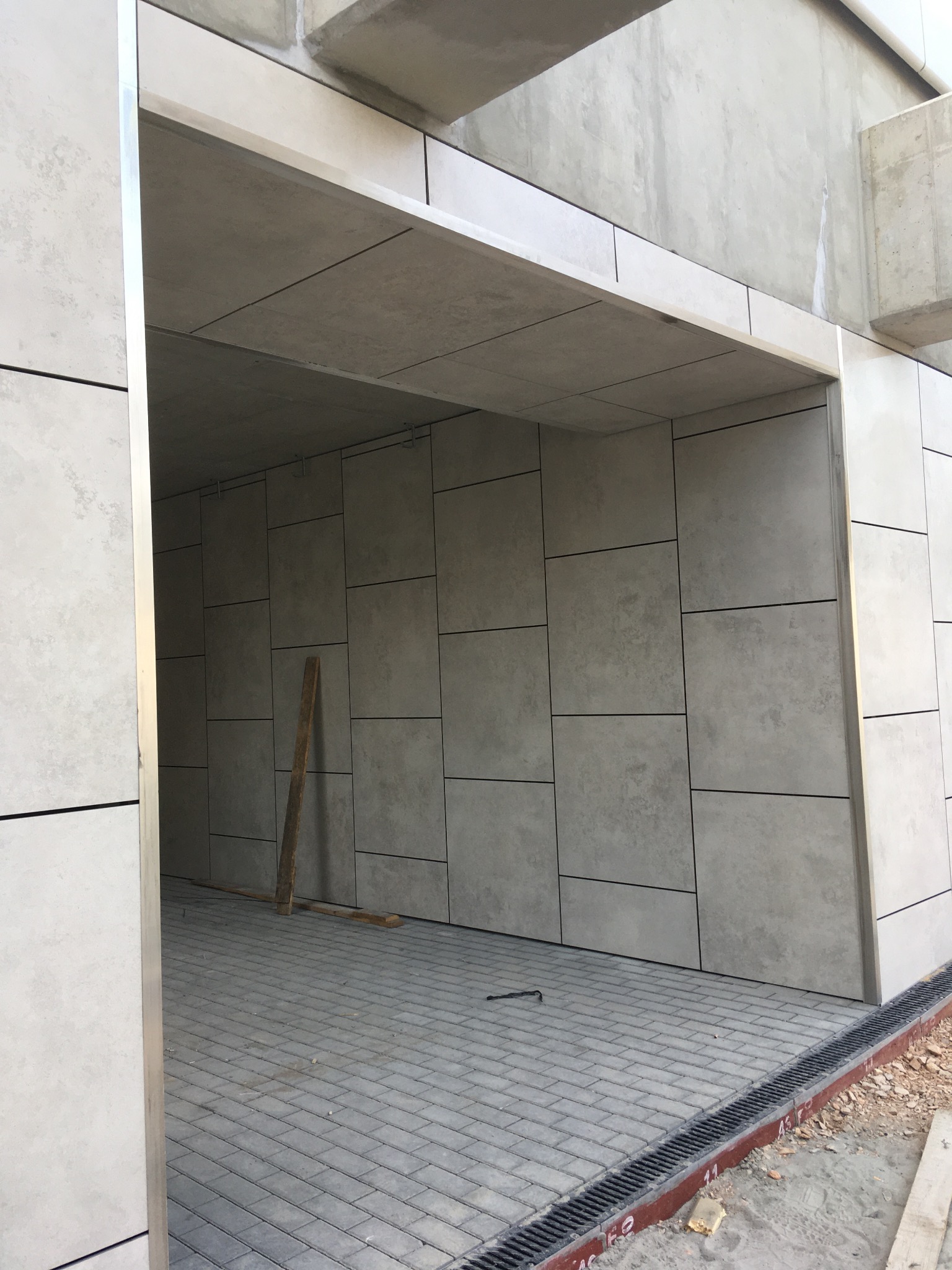 Wykończone wejście do budynku z szarą elewacją z płyt, widoczna kostka brukowa i odpływ liniowy. Elementy wykończeniowe z metalu. Lekkie oświetlenie.