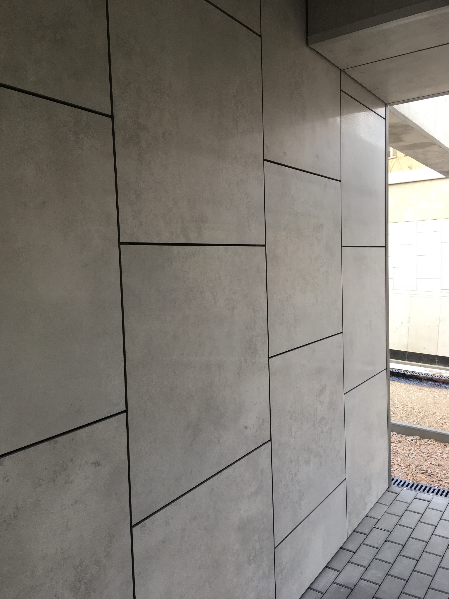 Elewacja budynku wykończona szarymi panelami z ciemnymi fugami, widok z boku, z fragmentem chodnika z kostki brukowej i odpływem liniowym w tle.