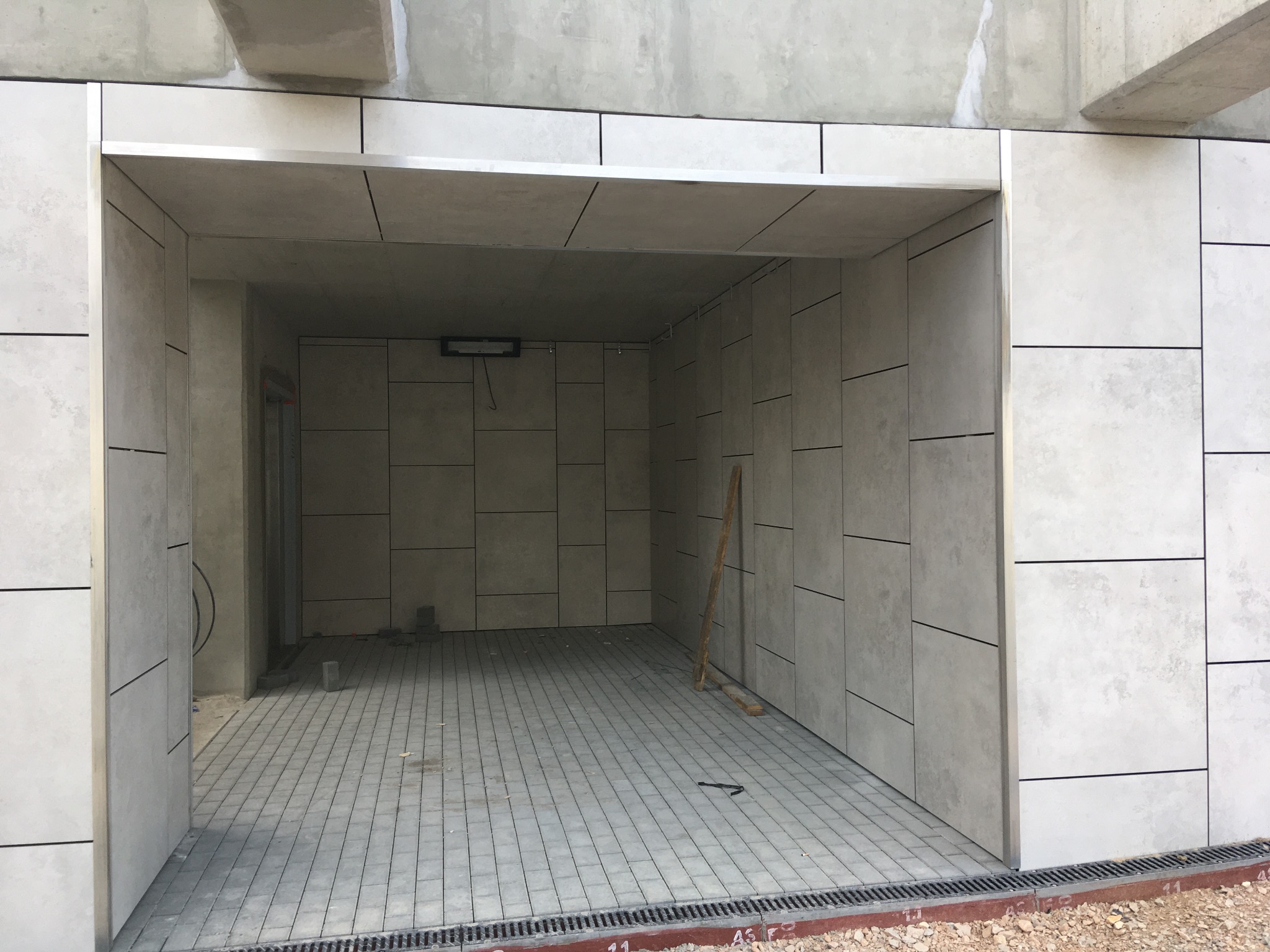 Wykończone wejście do garażu z szarymi płytami elewacyjnymi i kostką brukową. Widoczne detale wykończeniowe i drenaż. Minimalistyczny design.