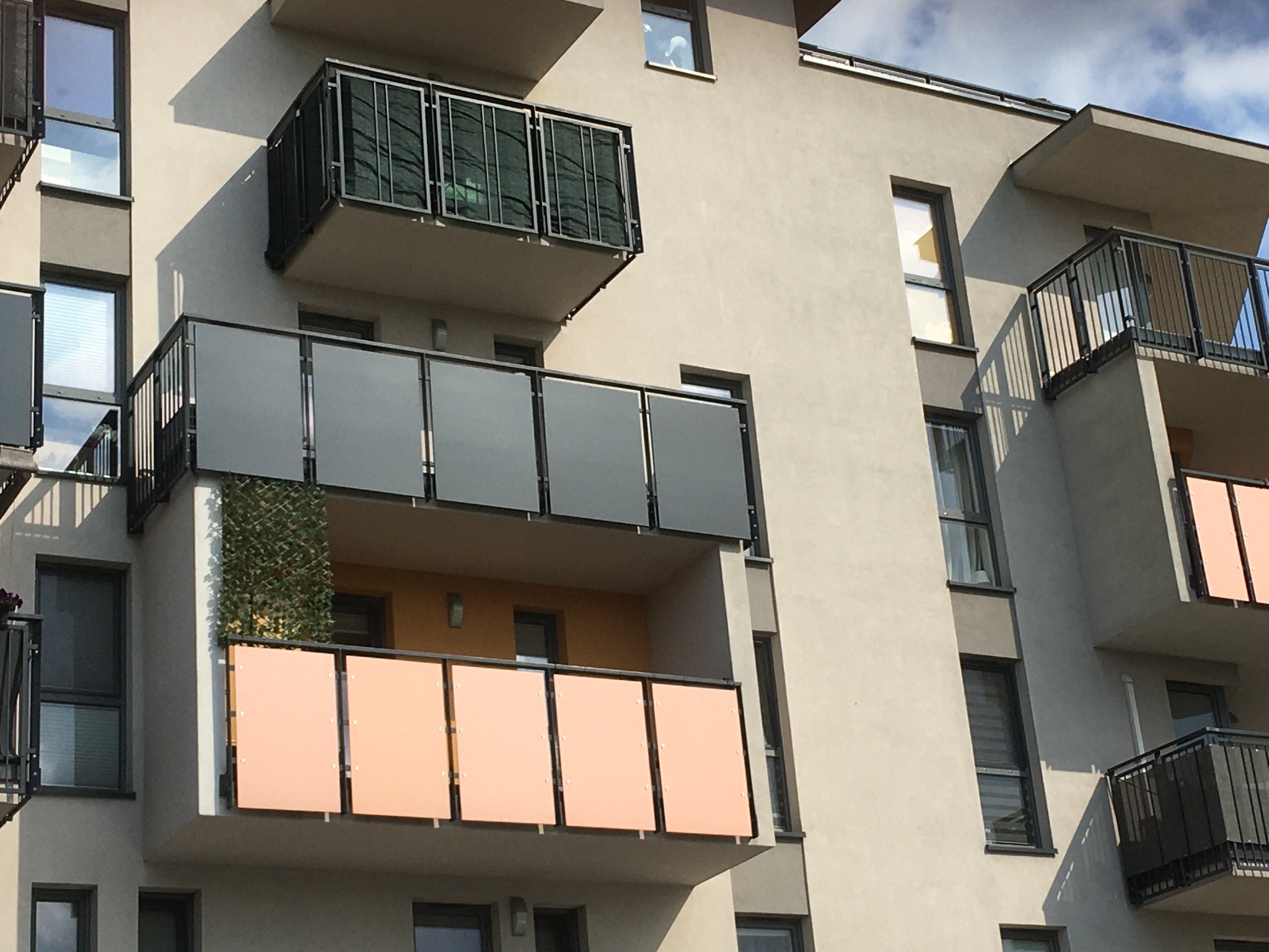 Nowoczesny blok z elewacją w odcieniach szarości i beżu. Balkony z różnymi wypełnieniami: szkło, metalowa siatka i panele w kolorze brzoskwini.