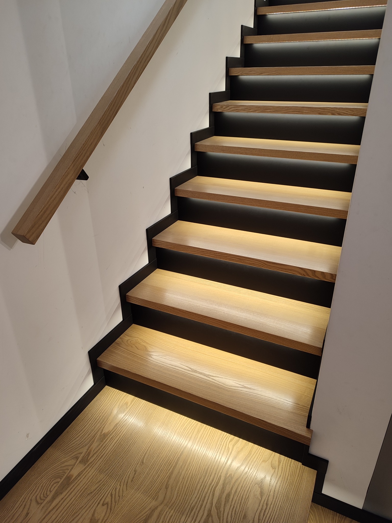 Nowoczesne schody z drewna o jasnym odcieniu, z czarnymi elementami konstrukcyjnymi i podświetleniem LED pod każdym stopniem, widoczne z góry pod kątem.