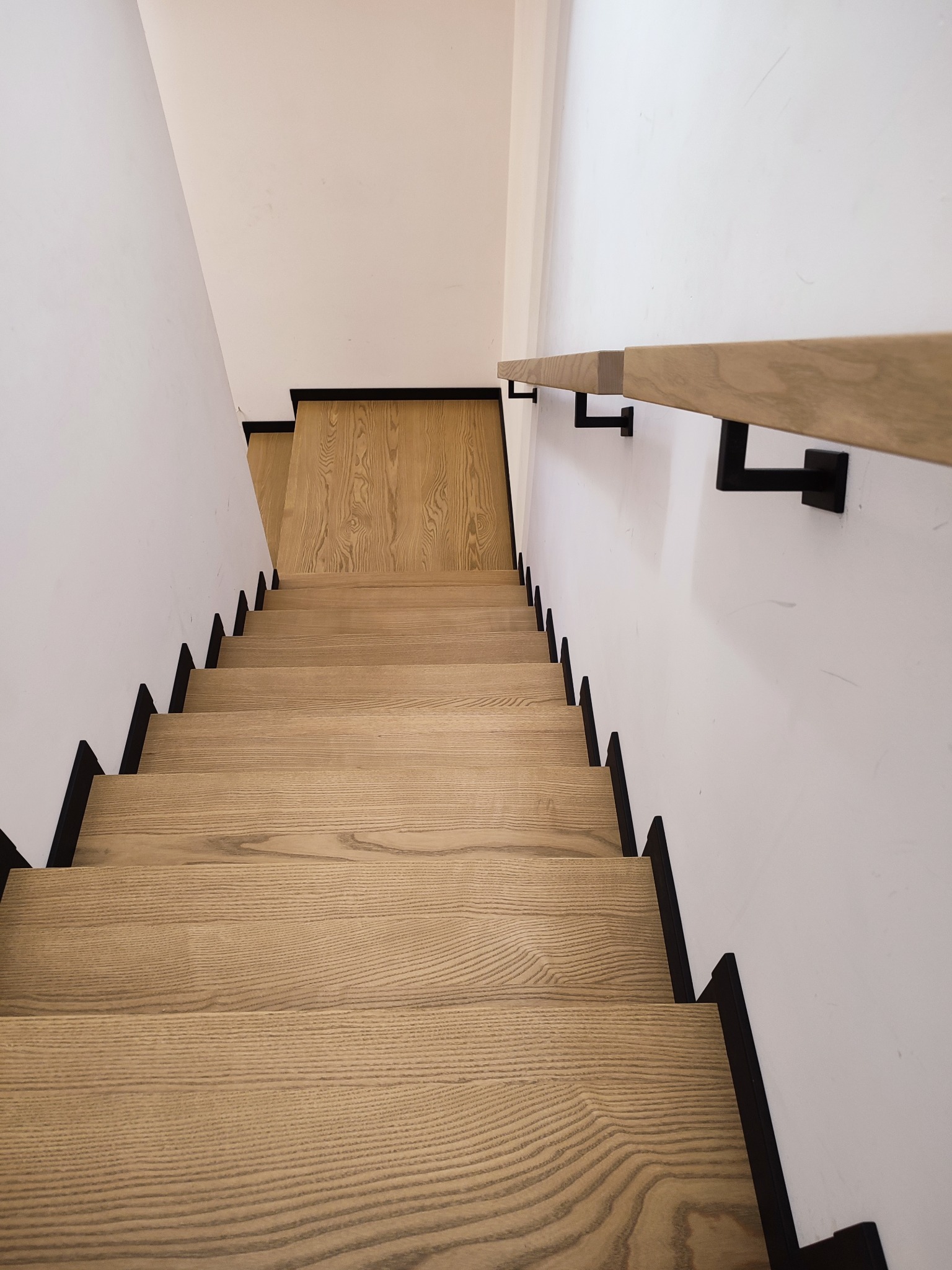 Widok z góry na jasne, drewniane schody z czarnymi podstopnicami i prostą, drewnianą poręczą zamocowaną na czarnych wspornikach.