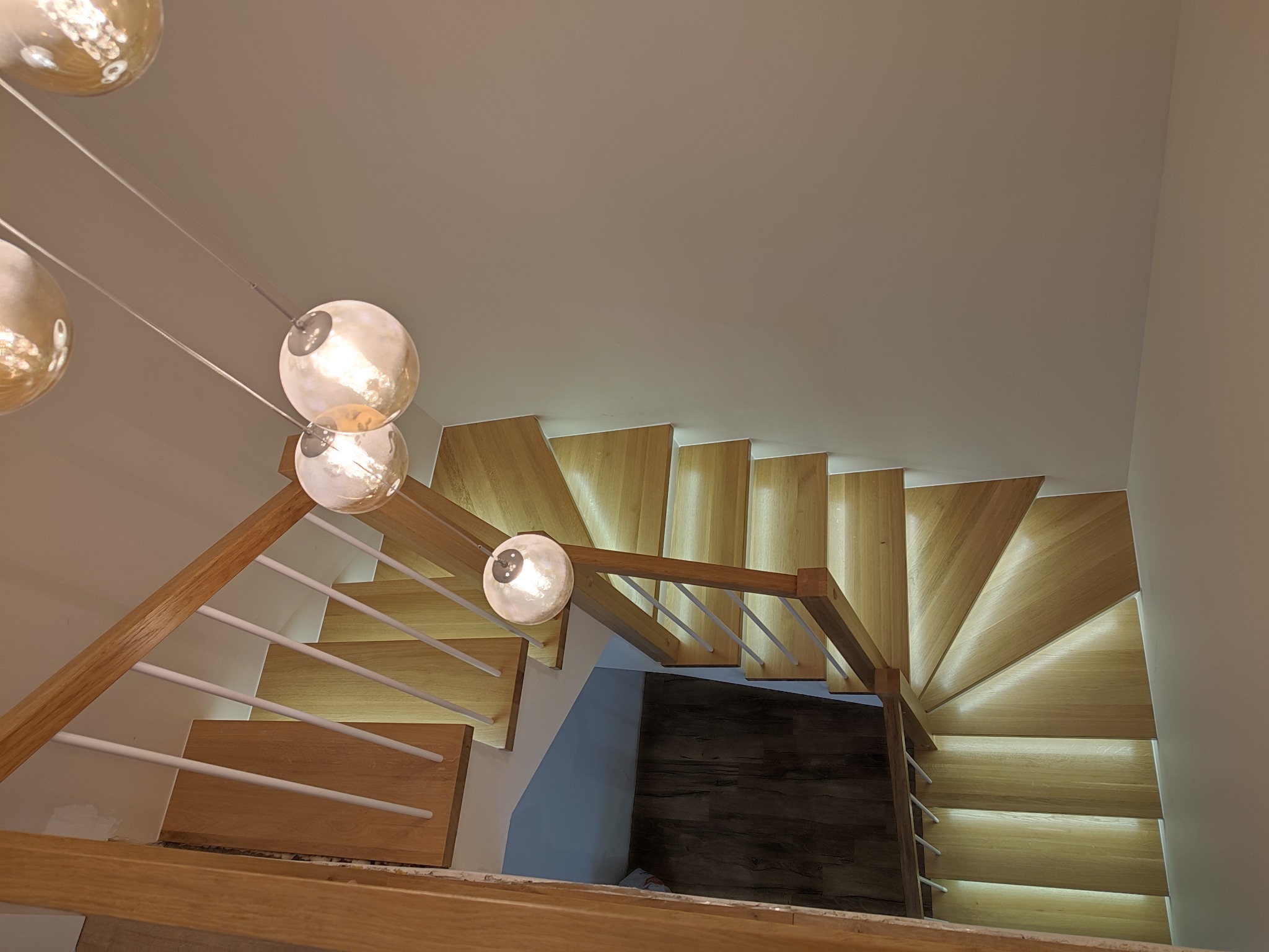 Widok z góry na jasne, drewniane schody spiralne z białymi balustradami, oświetlone wiszącymi lampami z kulistymi kloszami.