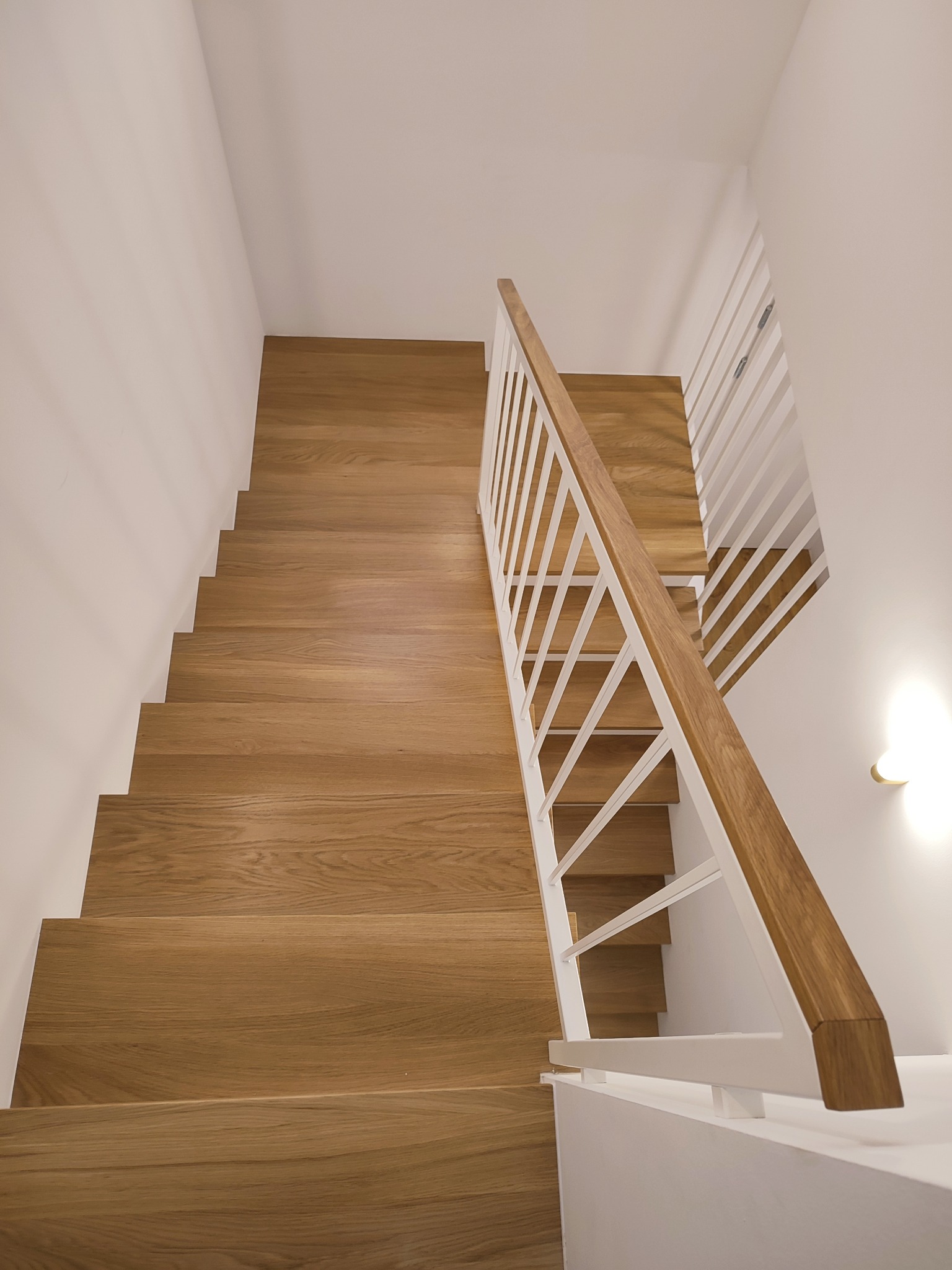 Widok z góry na drewniane schody z jasną balustradą z białymi, pionowymi elementami i drewnianym poręczą, oświetlone przez ciepłe światło kinkietu na białej ścianie.