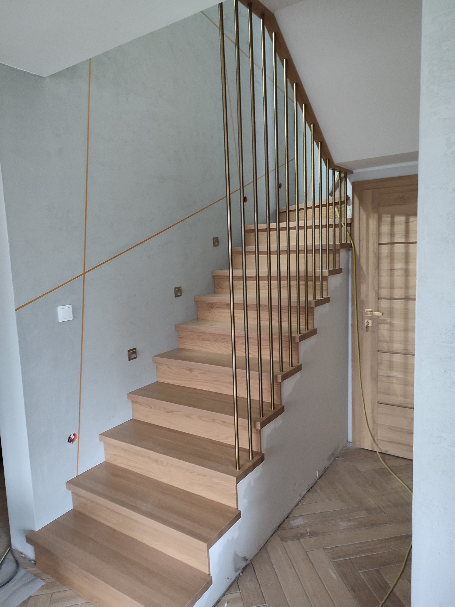 Jasne, drewniane schody z minimalistyczną, złotą balustradą w nowoczesnym wnętrzu z dekoracyjną ścianą.