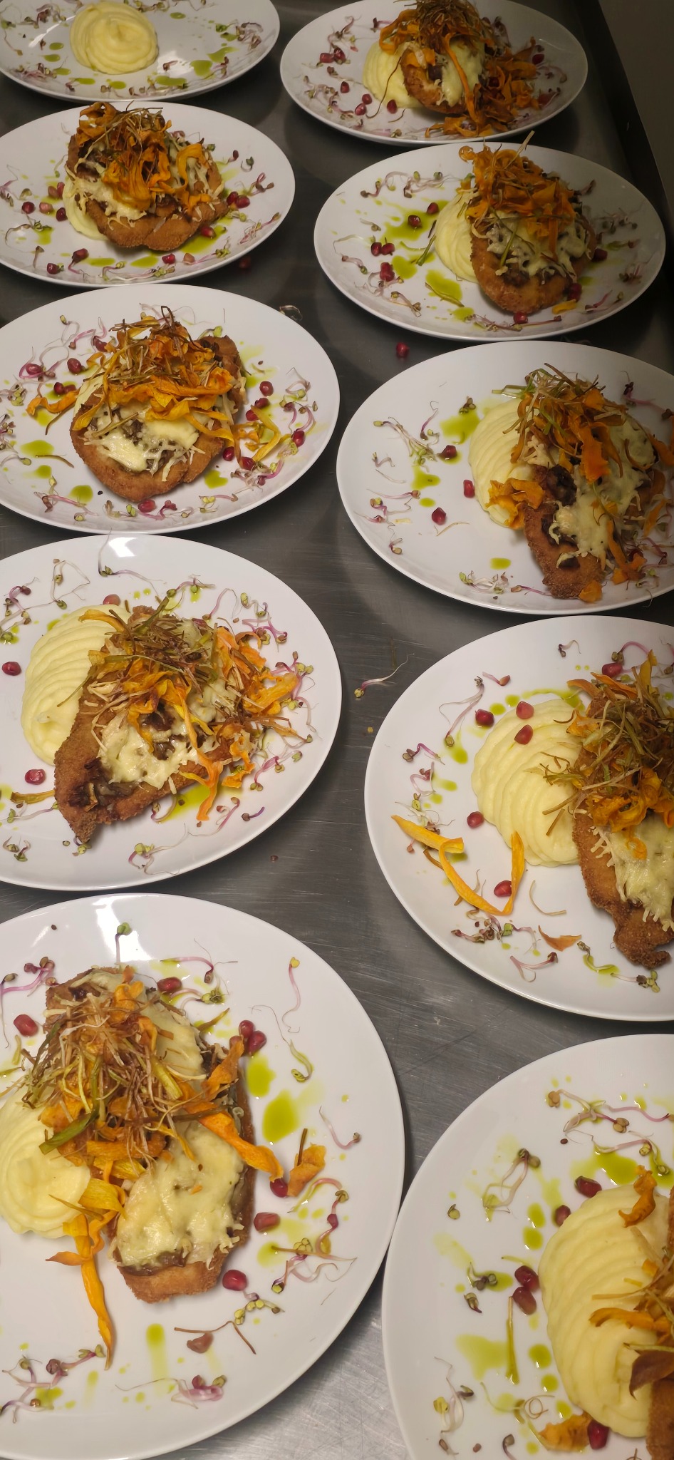 Rzędy talerzy z panierowanym mięsem, puree i dekoracjami z kiełków, granatu i oleju ziołowego. Elegancki catering na metalowym stole.