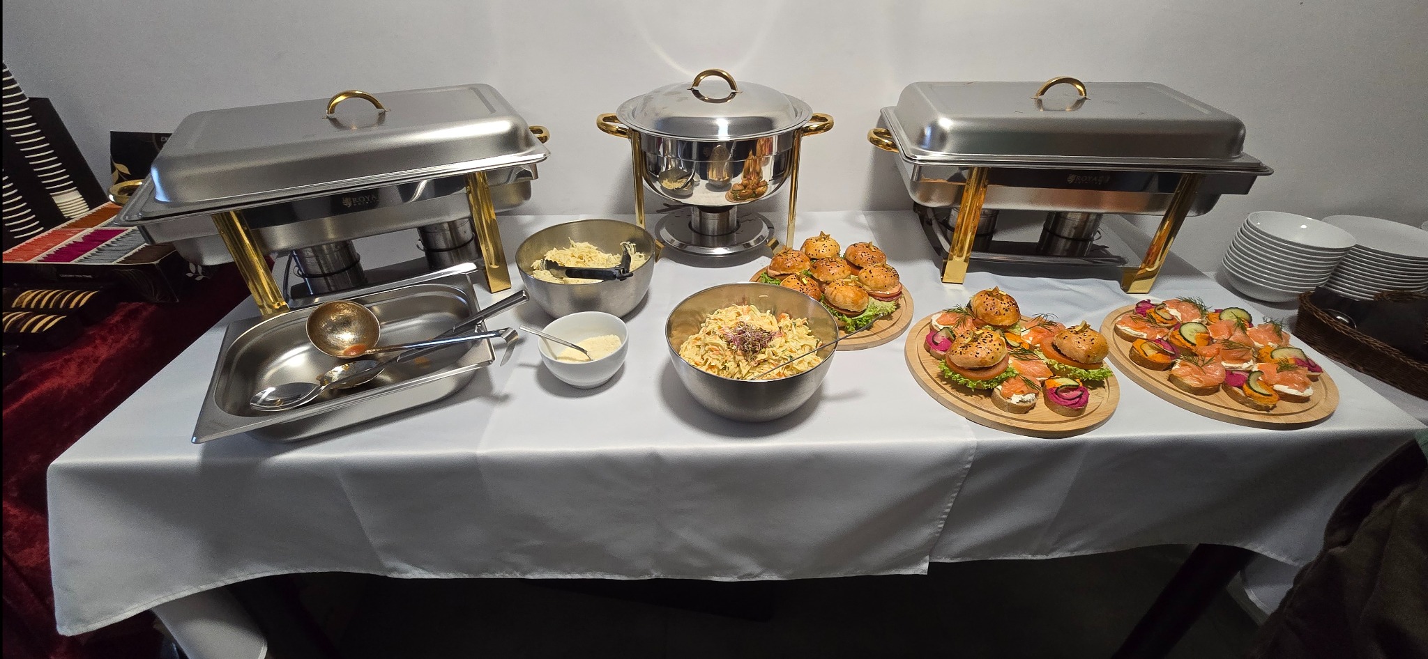 Stół cateringowy z podgrzewaczami, sałatkami i mini-kanapeczkami z łososiem. Bufet szwedzki na białym obrusie, widok z góry, oświetlenie studyjne.