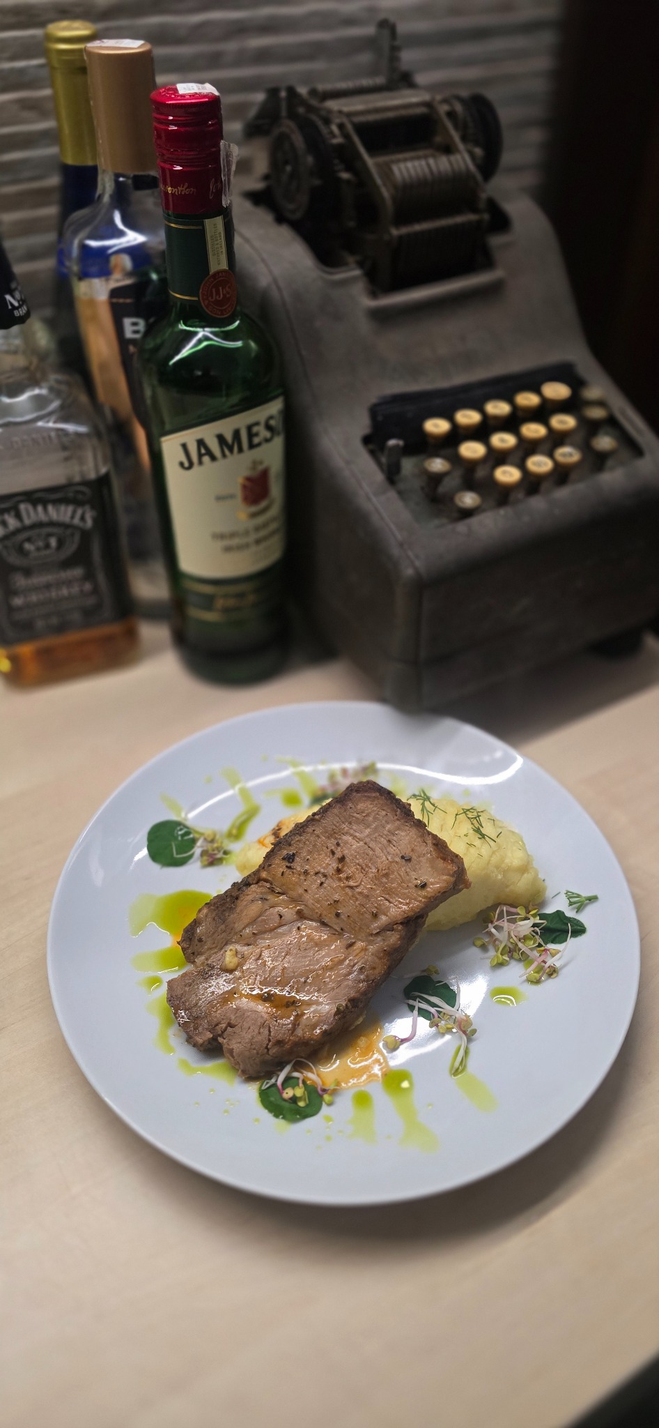Wyjątkowe danie mięsne z puree i dekoracjami na talerzu, w tle butelki alkoholu i zabytkowa kasa fiskalna. Catering w Tarnowie z nutą retro.