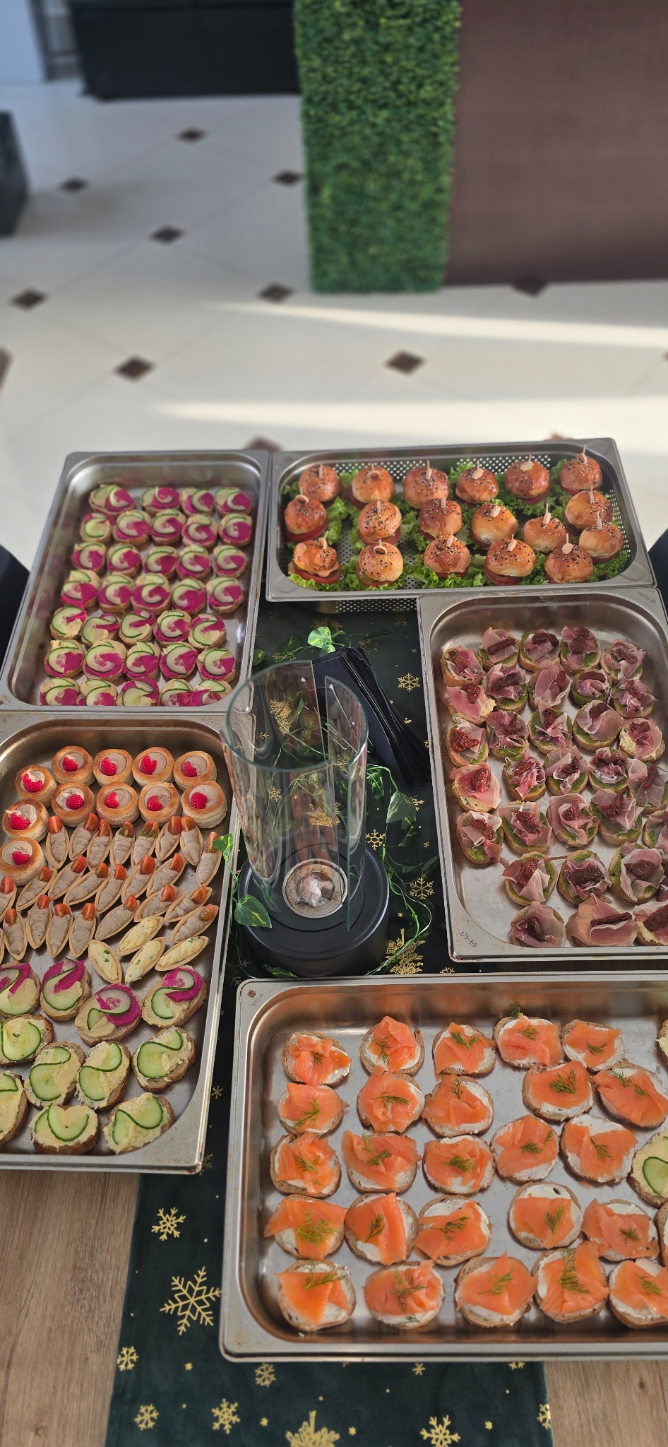 Stół z różnorodnymi przekąskami w metalowych tacach: mini burgery, kanapki z łososiem, figami i szynką, tartaletki. Dekoracja świąteczna w tle.