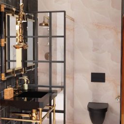 MAP Projekt - Ekskluzywna łazienka z czarną umywalką na złotym stelażu, kabiną prysznicową w stylu loft i podwieszaną toaletą na tle marmurowej ściany. Złote detale dodają elegancji.