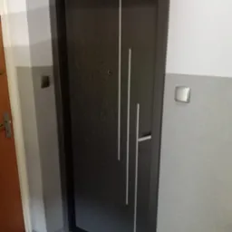 Ciemne, nowoczesne drzwi z trzema pionowymi, białymi paskami, zamontowane w szarym korytarzu z widocznym włącznikiem światła. Widok z bliska, z boku.