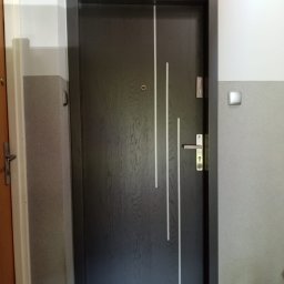 MAGNUM URSZULA GAWROŃSKA - Ciemnoszare drzwi wejściowe z trzema pionowymi, białymi paskami po prawej stronie, zamontowane w szarym korytarzu, z widocznym wizjerem i klamką.