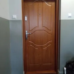 Brązowe drzwi wejściowe wewnątrz budynku z szarą ścianą i widocznym zamkiem oraz wizjerem. Drzwi posiadają ozdobne frezy i srebrną klamkę.