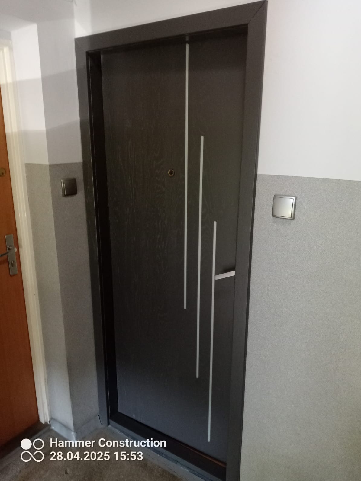 Ciemne, nowoczesne drzwi z trzema pionowymi, białymi paskami, zamontowane w szarym korytarzu z widocznym włącznikiem światła. Widok z bliska, z boku.