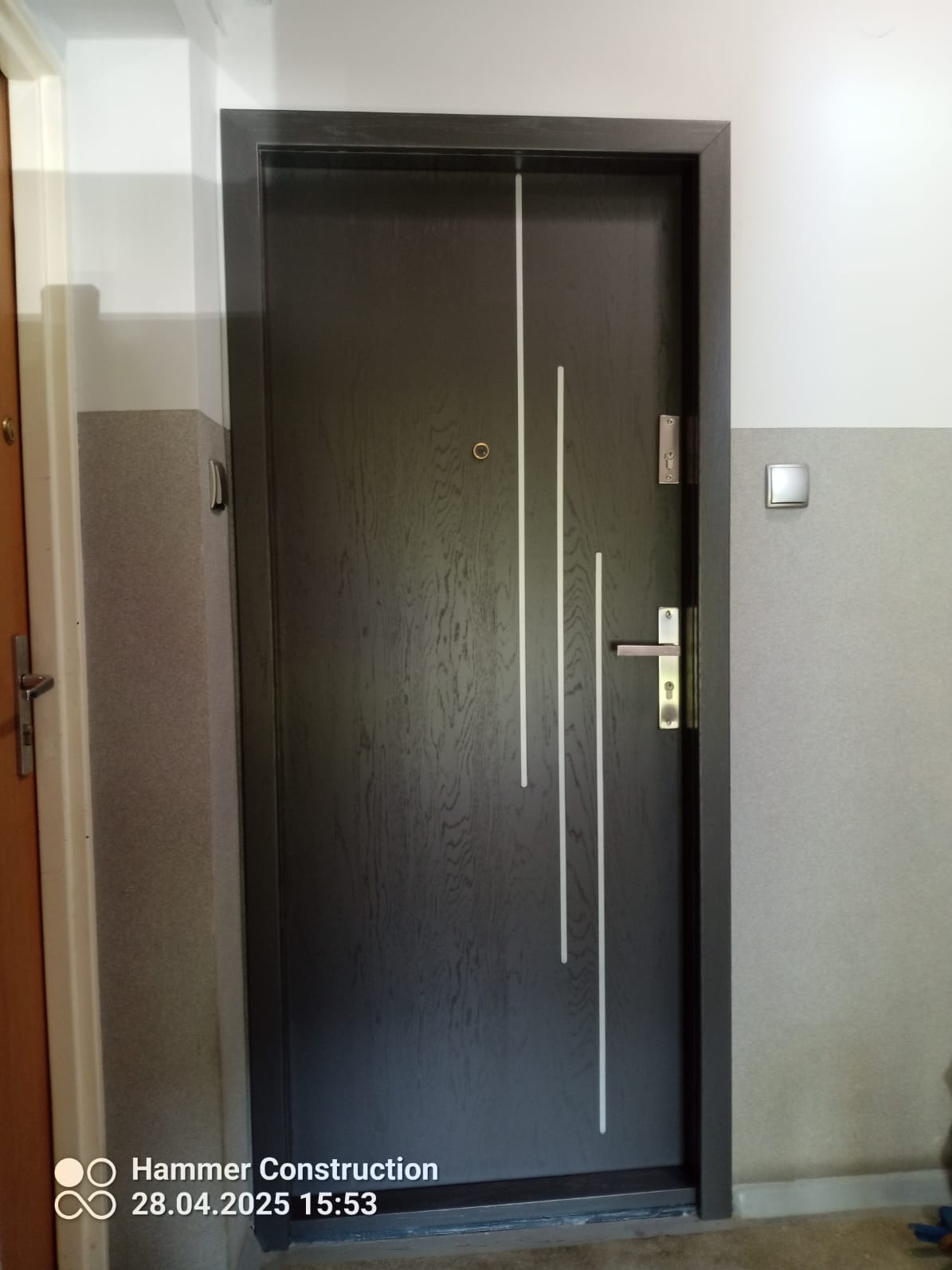 Ciemnoszare drzwi wejściowe z trzema pionowymi, białymi paskami po prawej stronie, zamontowane w szarym korytarzu, z widocznym wizjerem i klamką.