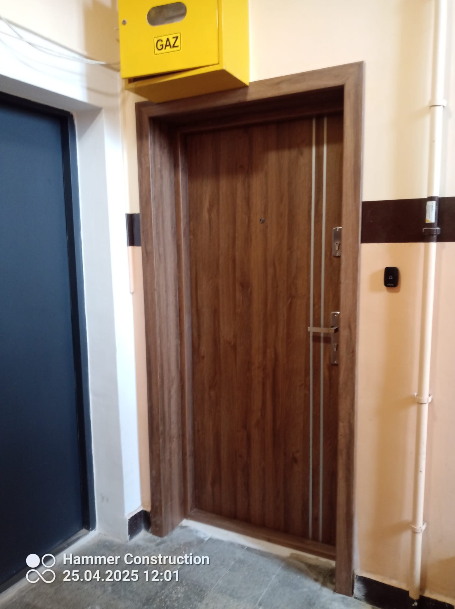 Brązowe drzwi z trzema pionowymi, jasnymi paskami w Oleśnicy. Nad nimi żółta skrzynka gazowa. Widoczny fragment ciemnych drzwi po lewej stronie.