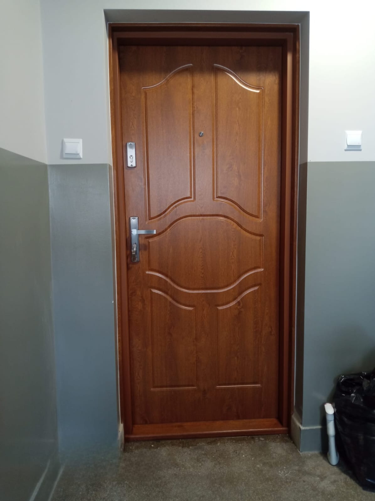 Brązowe drzwi wejściowe wewnątrz budynku z szarą ścianą i widocznym zamkiem oraz wizjerem. Drzwi posiadają ozdobne frezy i srebrną klamkę.