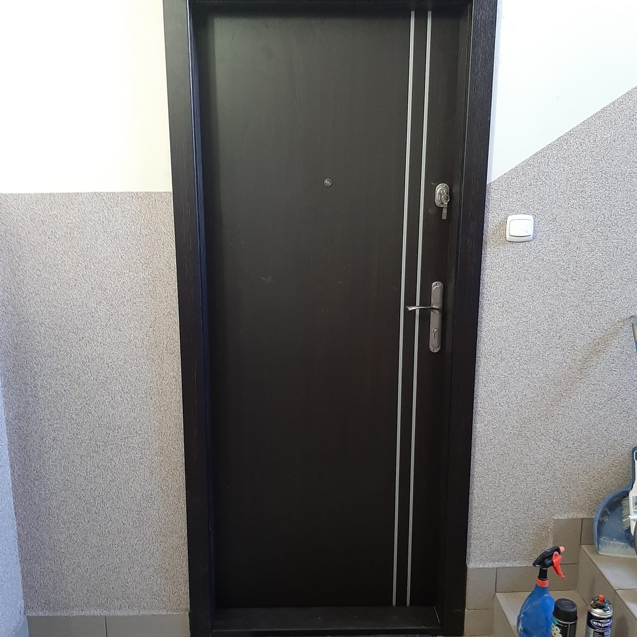Ciemne drzwi wejściowe z dwoma pionowymi, jasnymi paskami, zamontowane w korytarzu. Widoczny zamek z kluczem, wizjer i klamka. W tle ściana z szarą tapetą strukturalną.