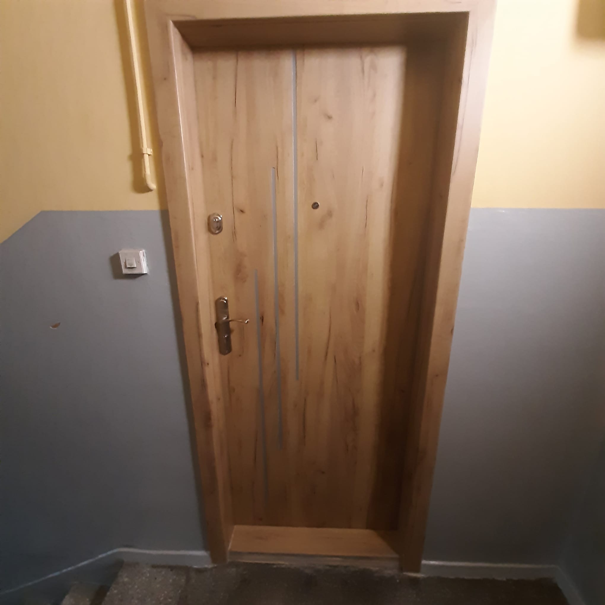 Drewniane drzwi wejściowe z metalowymi akcentami, zamontowane w klatce schodowej z żółto-szarymi ścianami. Widoczne elementy wykończenia i zamek.