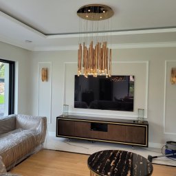 DAN-HOUSE SPÓŁKA Z OGRANICZONĄ ODPOWIEDZIALNOŚCIĄ - Salon po remoncie z nowoczesnym oświetleniem: wisząca lampa z złotych rurek, złote kinkiety, telewizor na ścianie, meble z drewnianymi frontami, marmurowa podłoga, sofa zabezpieczona folią...