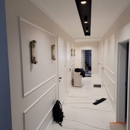 DAN-HOUSE SPÓŁKA Z OGRANICZONĄ ODPOWIEDZIALNOŚCIĄ - Nowoczesny korytarz po remoncie z białymi ścianami zdobionymi sztukaterią, marmurową podłogą, czarnym oświetleniem sufitowym i złotymi lampami ściennymi, w tle widoczne schody.