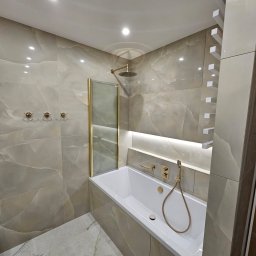 DAN-HOUSE SPÓŁKA Z OGRANICZONĄ ODPOWIEDZIALNOŚCIĄ - Wykończona łazienka z wanną obudowaną płytkami imitującymi marmur, złotą armaturą i deszczownicą za szklanym panelem.