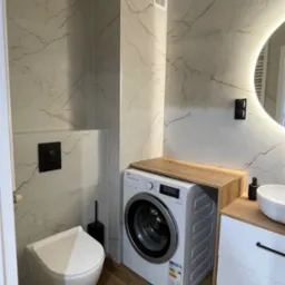 Nowoczesna łazienka z płytkami imitującymi marmur, wiszącą toaletą i pralką zabudowaną drewnianym blatem. Okrągłe lustro z podświetleniem, drewniana podłoga.