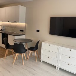 DAN-HOUSE SPÓŁKA Z OGRANICZONĄ ODPOWIEDZIALNOŚCIĄ - Nowoczesna aranżacja wnętrza: biała zabudowa kuchenna z marmurowym akcentem, czarne krzesła przy okrągłym stole i biała komoda pod telewizorem. Jasna podłoga.