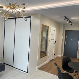 DAN-HOUSE SPÓŁKA Z OGRANICZONĄ ODPOWIEDZIALNOŚCIĄ - Nowoczesne wnętrze mieszkania: biała szafa w zabudowie, złoty żyrandol, lustro w zdobionej ramie i minimalistyczne meble w neutralnej kolorystyce.